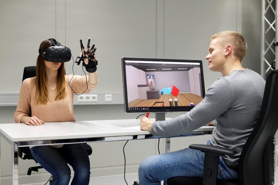 A woman wearing a virtual reality (VR) headset and motion-tracking gloves interacts with a virtual environment, while a man sitting across from her observes the simulation on a computer screen. The screen displays a 3D-rendered room with interactive objects.